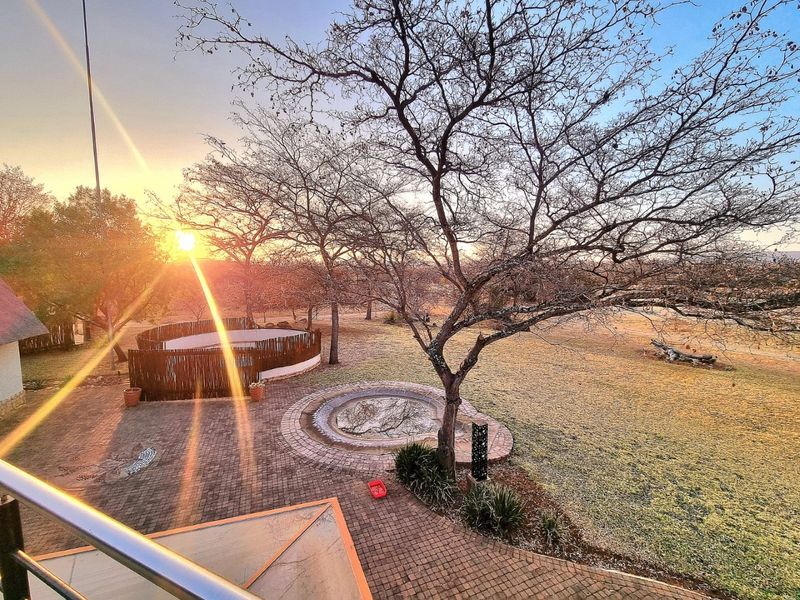 A scenic rear garden view featuring a paved patio, a circular water feature, and a boma area, captured at sunset from an elevated balcony.