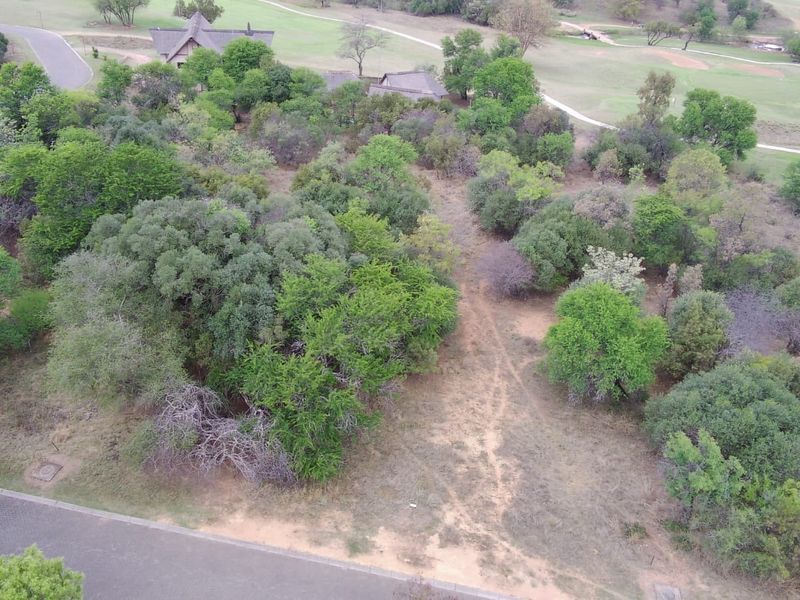 Aerial view of a vacant residential plot featuring established indigenous trees and direct access from a paved road within a golf estate setting.
