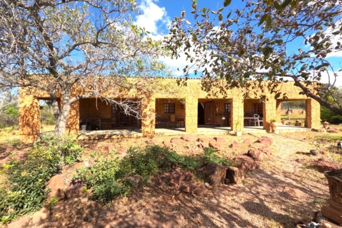 A rustic farm-style dwelling featuring a covered patio with stone pillars and masonry walls, set within a natural, rocky landscape.