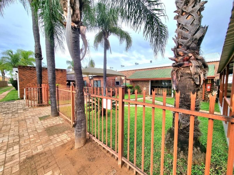 Rear exterior view of a commercial property featuring a brick facade, metal palisade fencing, a paved walkway, and a landscaped garden area.