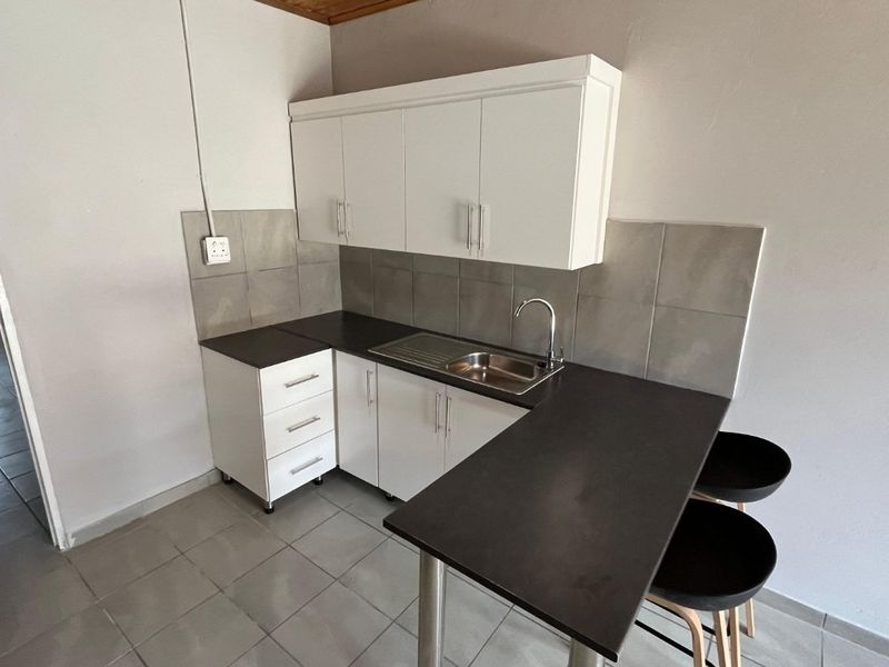 Compact kitchen featuring white built-in cabinetry, dark laminate countertops, stainless steel sink, and neutral tiled splashback with an integrated breakfast bar.