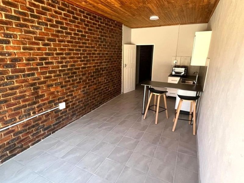 Open plan living area featuring an exposed brick feature wall, timber ceiling, and a compact kitchenette with built-in cabinetry and sink.