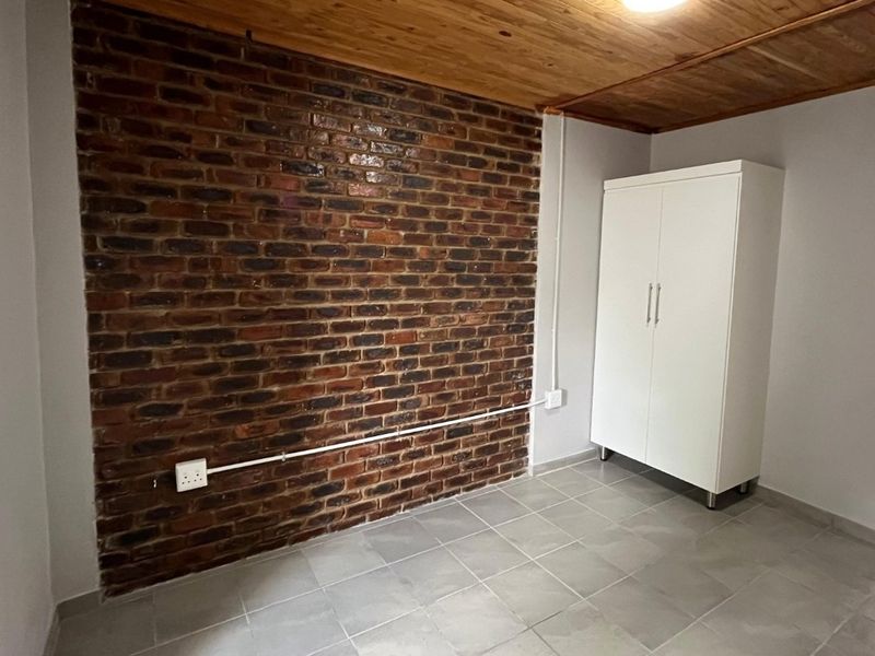 A bedroom featuring an exposed brick accent wall, timber ceiling, and neutral tiled flooring, offering a clean and functional space.