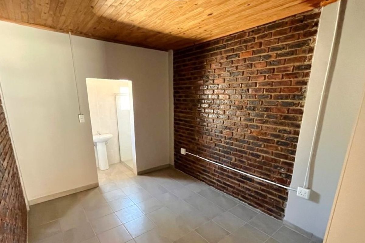 A bedroom featuring exposed brick walls, a timber ceiling, and tiled flooring, with an adjoining en-suite bathroom containing a pedestal basin and shower.