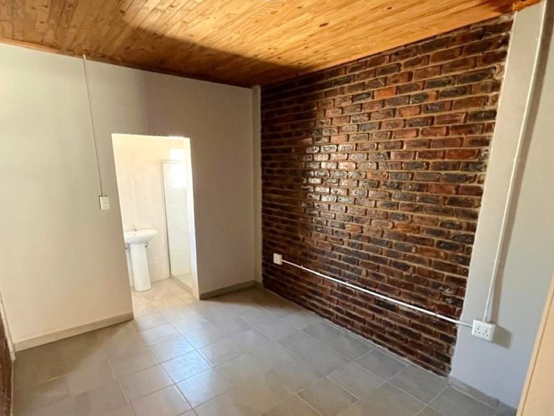 A bedroom featuring exposed brick walls, a timber ceiling, and tiled flooring, with an adjoining en-suite bathroom containing a pedestal basin and shower.
