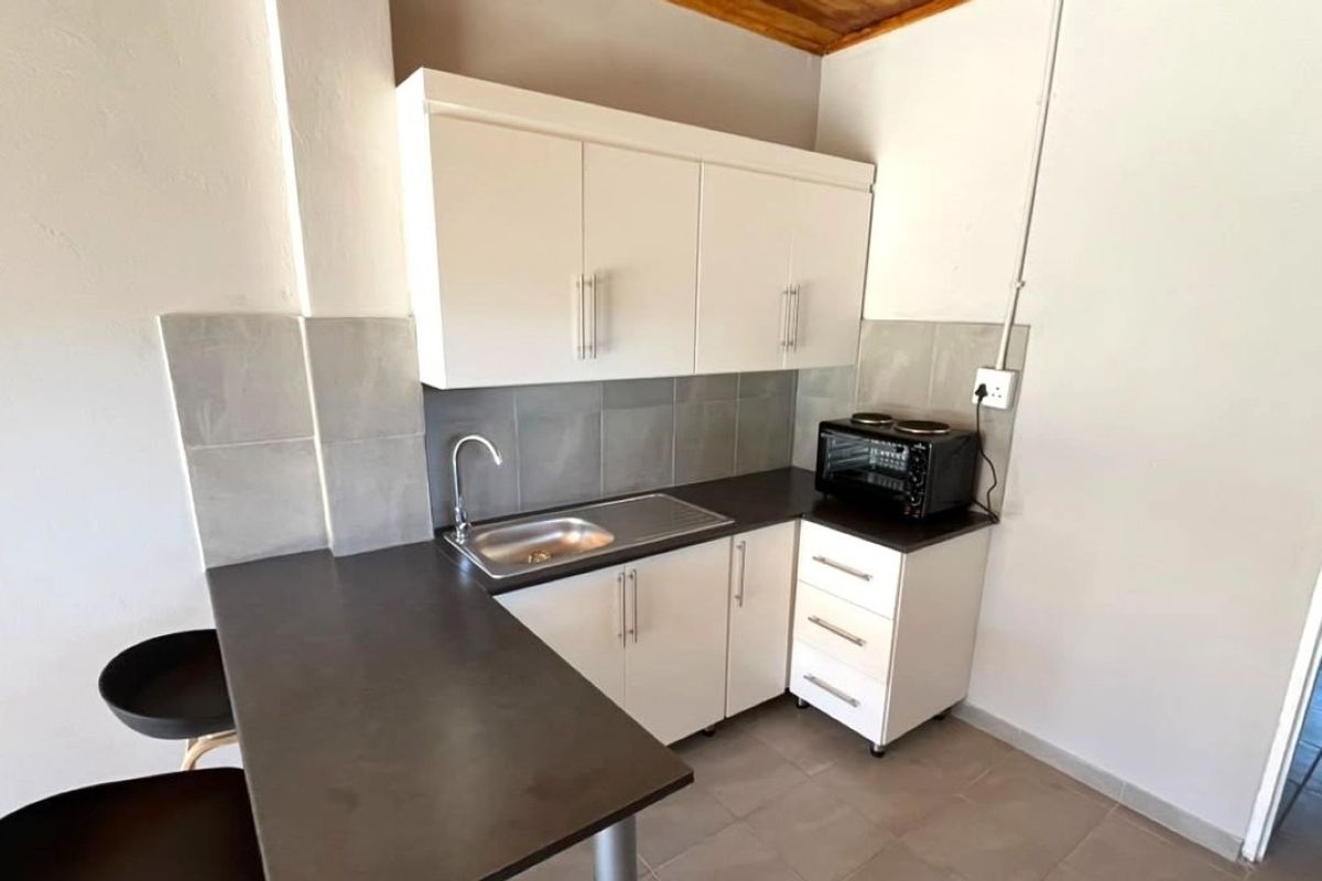 Compact kitchen featuring white built-in cabinetry, dark laminate countertops, a stainless steel sink, and neutral tiled backsplash and flooring.