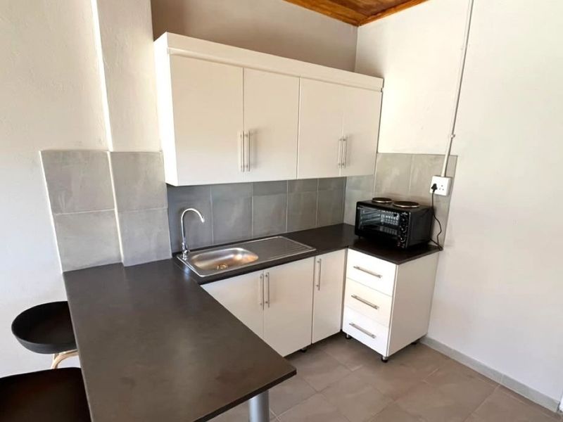 Compact kitchen featuring white built-in cabinetry, dark laminate countertops, a stainless steel sink, and neutral tiled backsplash and flooring.