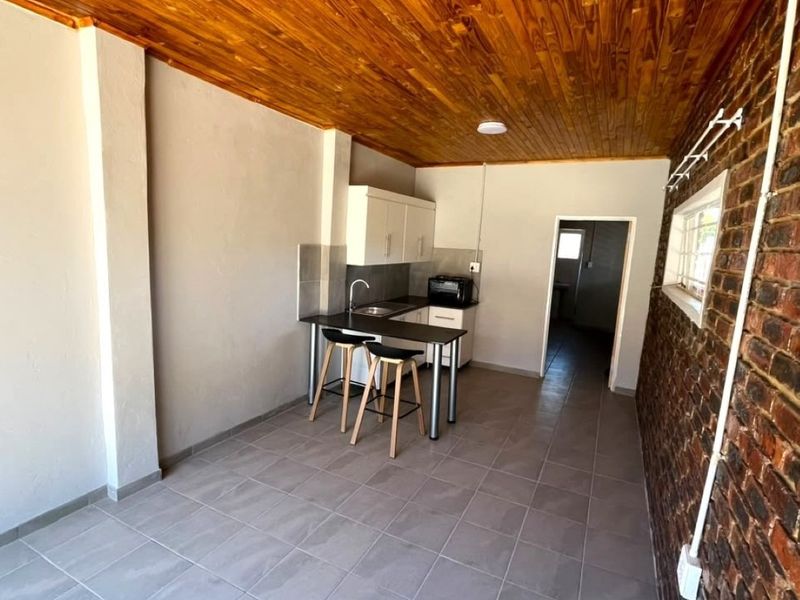 Open-plan living area featuring a rustic exposed brick wall, timber ceiling, tiled floors, and a compact kitchen with built-in cabinetry and a breakfast counter.