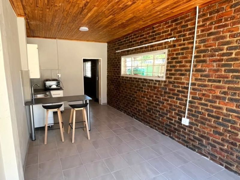 Open-plan living area featuring a rustic exposed brick wall, timber ceiling, tiled floors, and a compact kitchenette with built-in cabinetry.