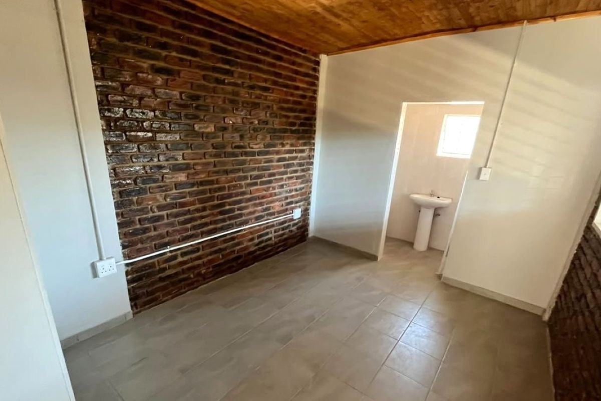 An empty bedroom featuring an exposed brick feature wall, wooden ceiling, tiled flooring, and an en-suite pedestal basin.