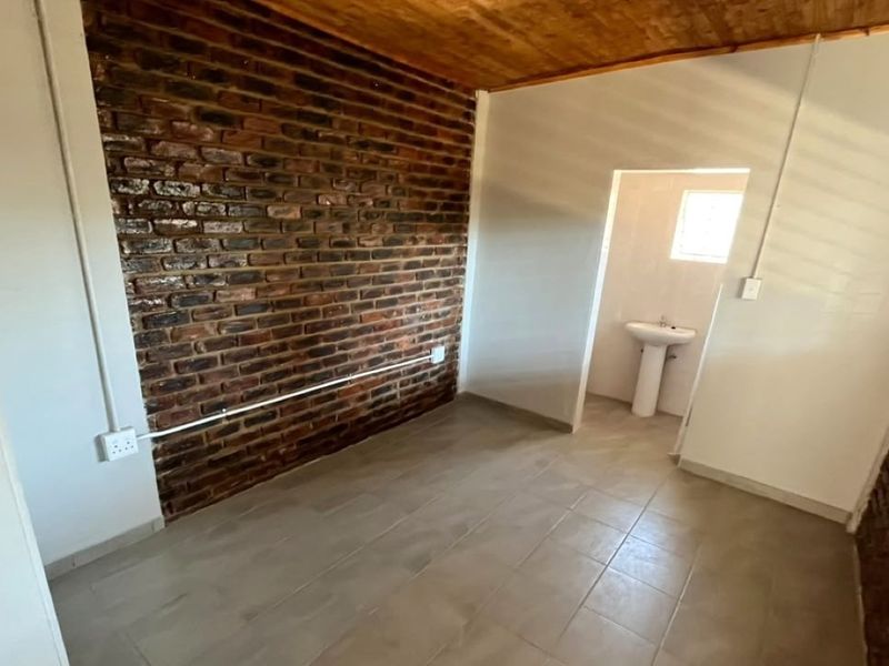 An empty bedroom featuring an exposed brick feature wall, wooden ceiling, tiled flooring, and an en-suite pedestal basin.