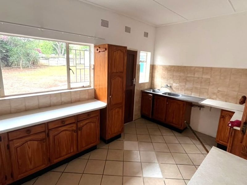 A functional kitchen featuring built-in wooden cabinetry, laminate countertops, a double stainless steel sink, and tiled flooring with large windows overlooking the garden.
