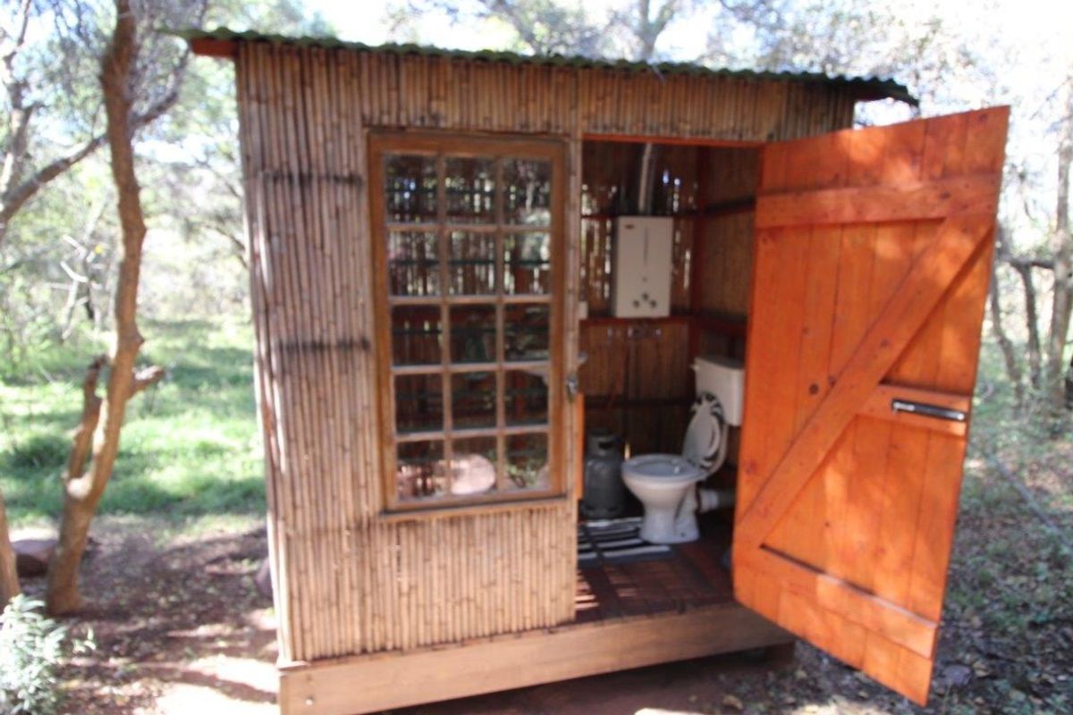 A rustic, off-grid outdoor bathroom structure featuring bamboo walls, a timber door, a toilet, and a gas water heater.