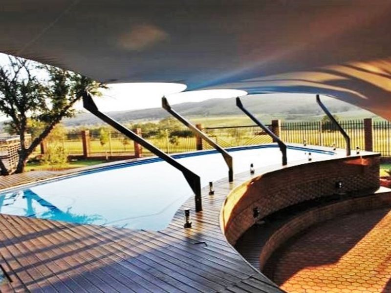 A well-maintained swimming pool area featuring a curved wooden deck, integrated brick seating, and a large shade sail, overlooking the surrounding farm landscape.