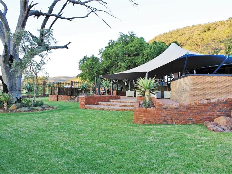 A well-maintained outdoor entertainment area featuring a large tensile shade structure, brick retaining walls, and landscaped garden surroundings on a farm property.
