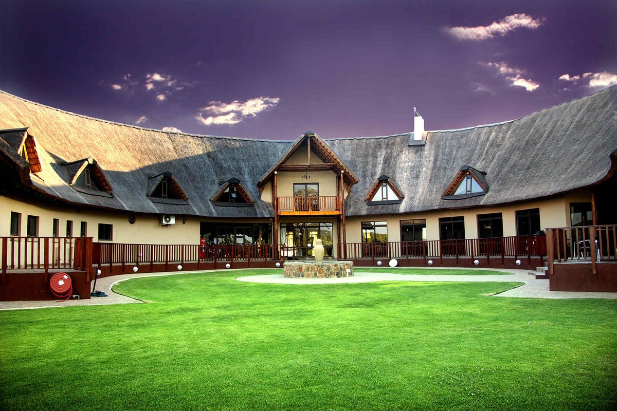 A spacious rear courtyard featuring a well-maintained lawn, a central stone fountain, and a large thatched-roof building with a balcony and wooden railings.