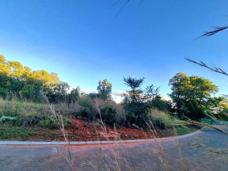 A view of a vacant residential plot featuring natural vegetation, sloping terrain, and paved street access.