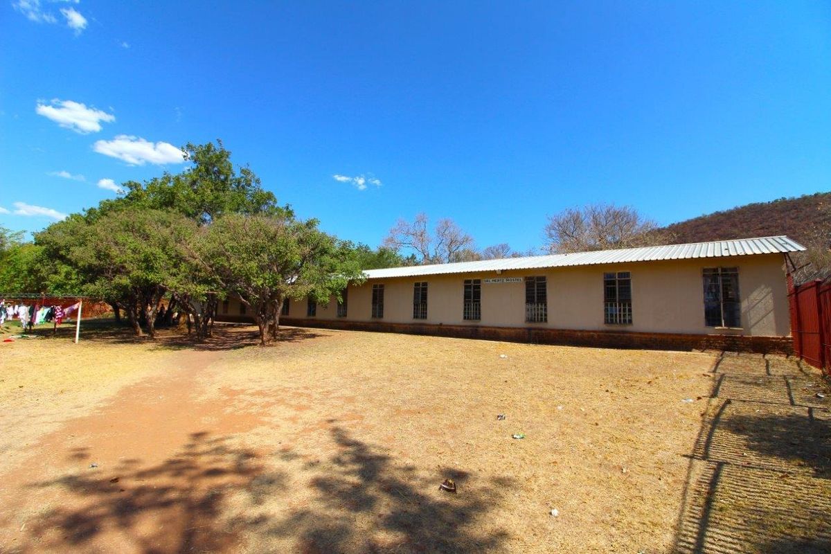 Exterior view of a long, single-story farm building with a metal roof, set within a large, open, and dry landscape.