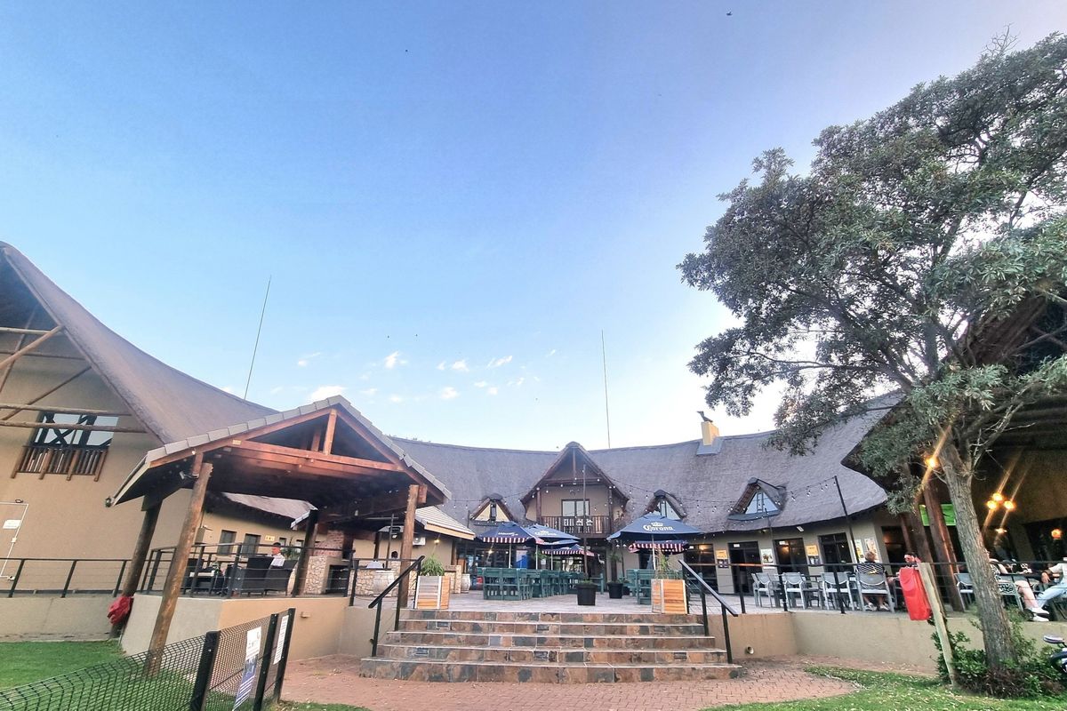 A spacious rear exterior view featuring a large thatched-roof building with stone stairs, paved areas, and covered patio spaces for outdoor entertainment.