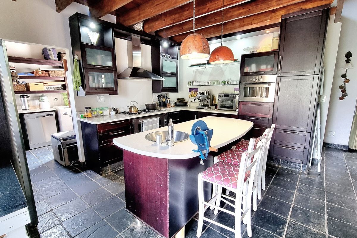 A spacious kitchen featuring dark wood cabinetry, a central island with a prep sink, stone countertops, and exposed timber ceiling beams.