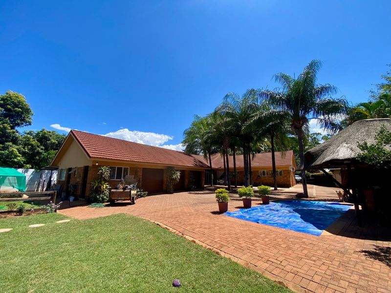 Spacious rear exterior featuring a brick-paved courtyard, swimming pool with cover, and a traditional thatched lapa, set against a well-maintained garden.