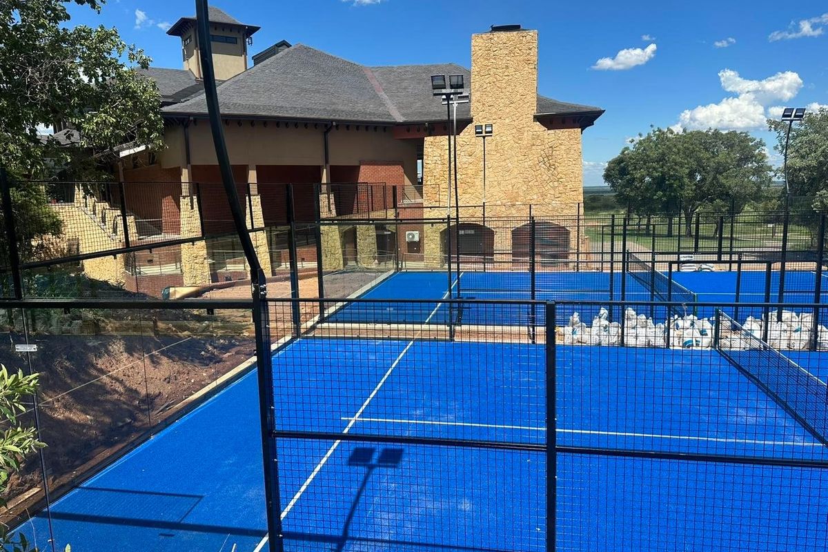 A well-maintained outdoor padel court facility featuring professional blue surfacing, perimeter fencing, and integrated floodlighting adjacent to a stone-clad building.