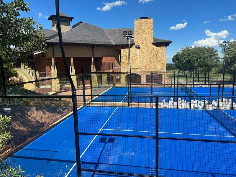 A well-maintained outdoor padel court facility featuring professional blue surfacing, perimeter fencing, and integrated floodlighting adjacent to a stone-clad building.