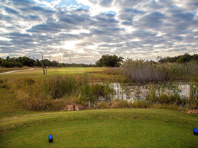 A scenic view of a golf course fairway featuring a natural pond and lush surrounding vegetation under a cloudy sky.