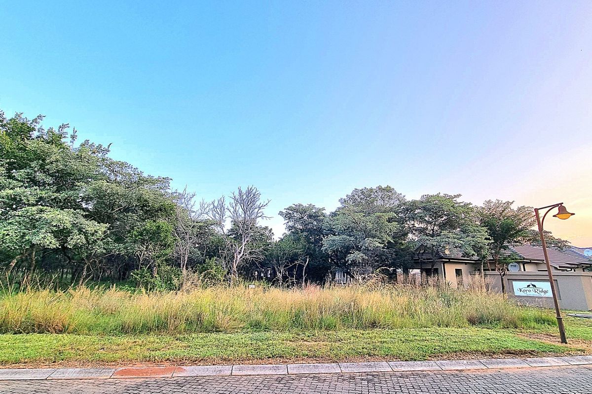 A vacant residential plot featuring established trees and natural vegetation, situated within a developed estate with paved street access.
