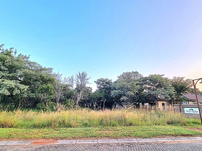 A vacant residential plot featuring established trees and natural vegetation, situated within a developed estate with paved street access.