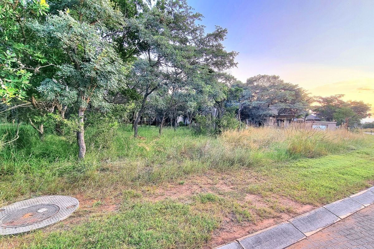 A view of a vacant residential plot featuring mature trees, natural grassland, and a paved curb along the street frontage.