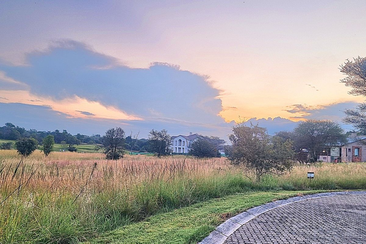 A scenic view of a vacant residential plot featuring natural grassland, a paved driveway edge, and distant estate homes under a sunset sky.