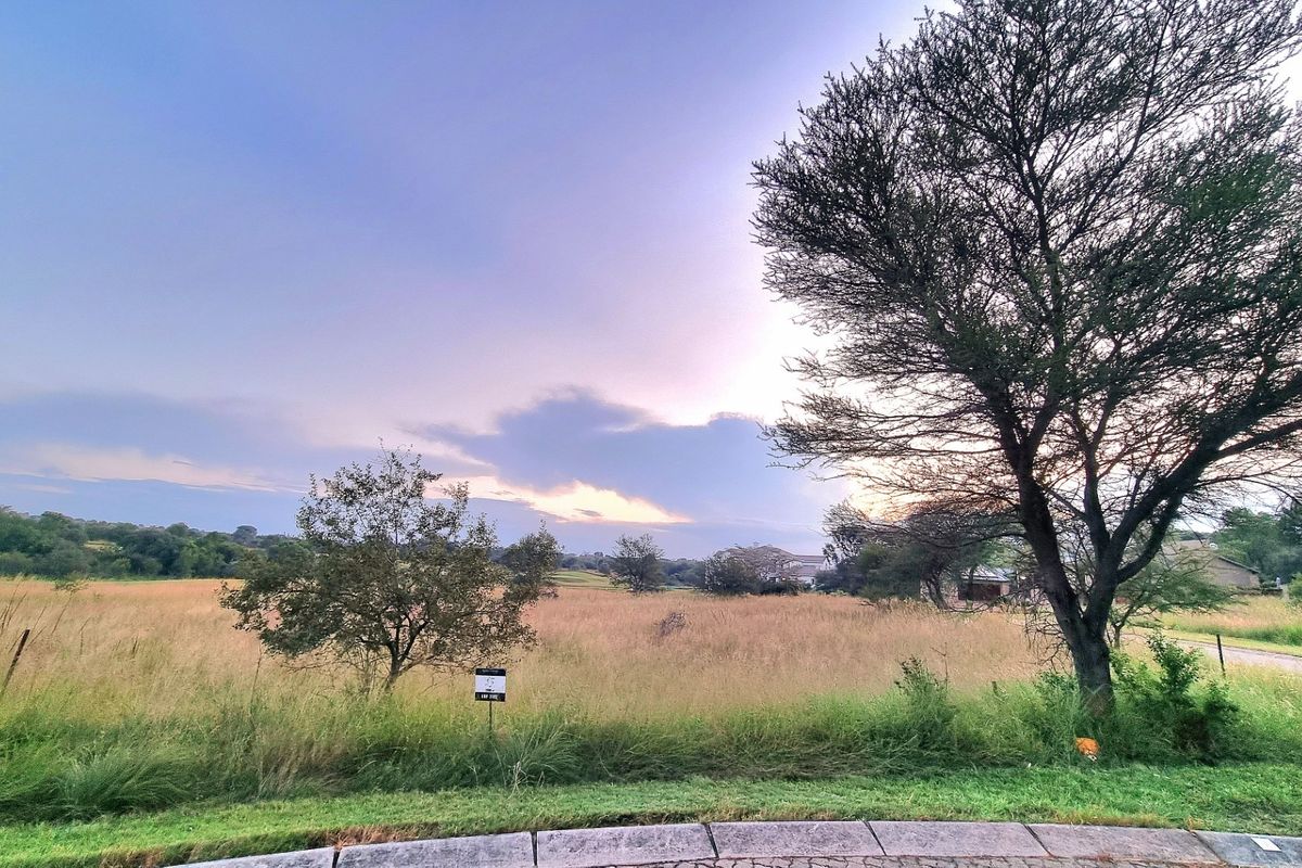 A view of a vacant residential plot featuring natural grass and mature trees, situated in a quiet neighborhood setting.
