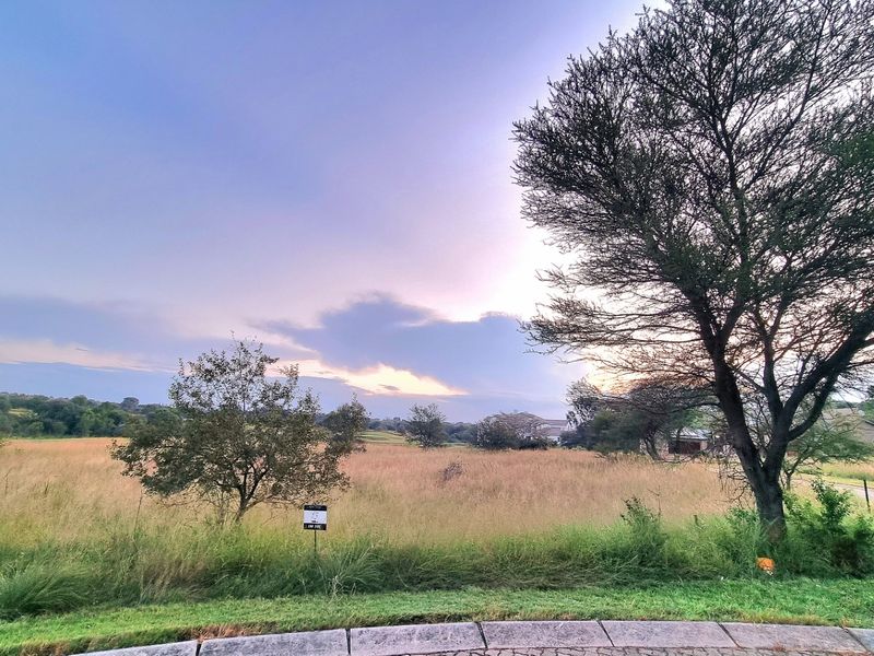 A view of a vacant residential plot featuring natural grass and mature trees, situated in a quiet neighborhood setting.