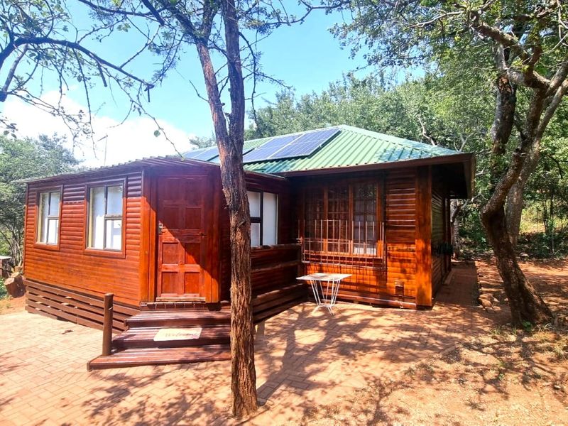 A charming timber log cabin featuring a corrugated metal roof with integrated solar panels, set on a paved patio area surrounded by mature trees.