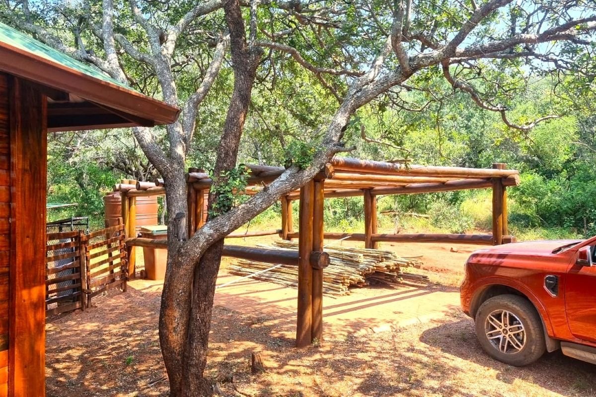 A rustic outdoor area featuring a timber pergola structure and wooden cabin cladding, set within a natural, wooded environment.