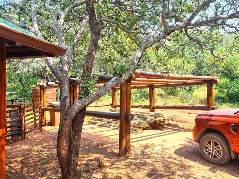 A rustic outdoor area featuring a timber pergola structure and wooden cabin cladding, set within a natural, wooded environment.
