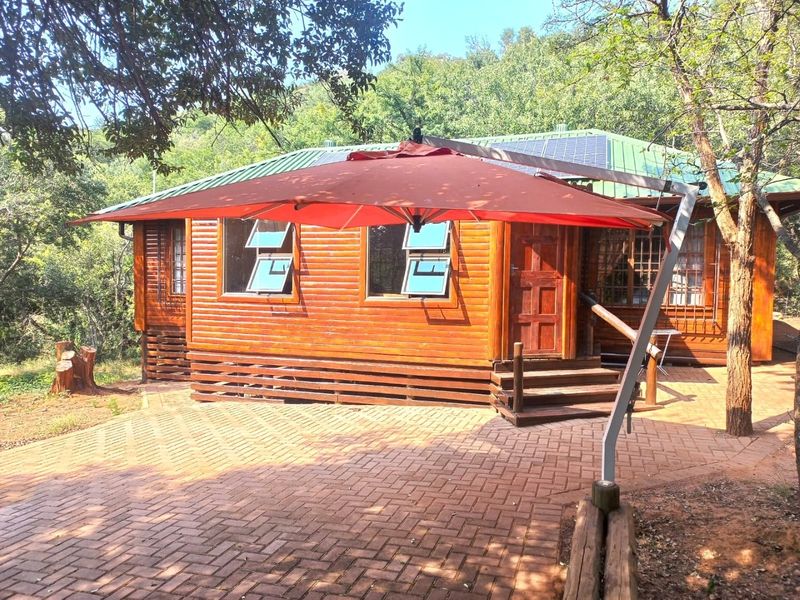 A charming timber log cabin featuring a paved entrance area, solar panels on the roof, and secure window fittings, set in a natural wooded environment.