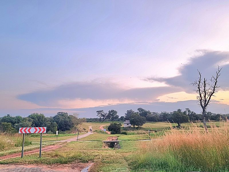 A view of a vacant residential plot featuring a dirt access road, natural grassland, and a small drainage culvert under a sunset sky.