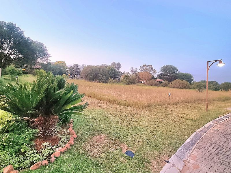 A view of a vacant residential plot featuring established landscaping, paved street access, and integrated street lighting.