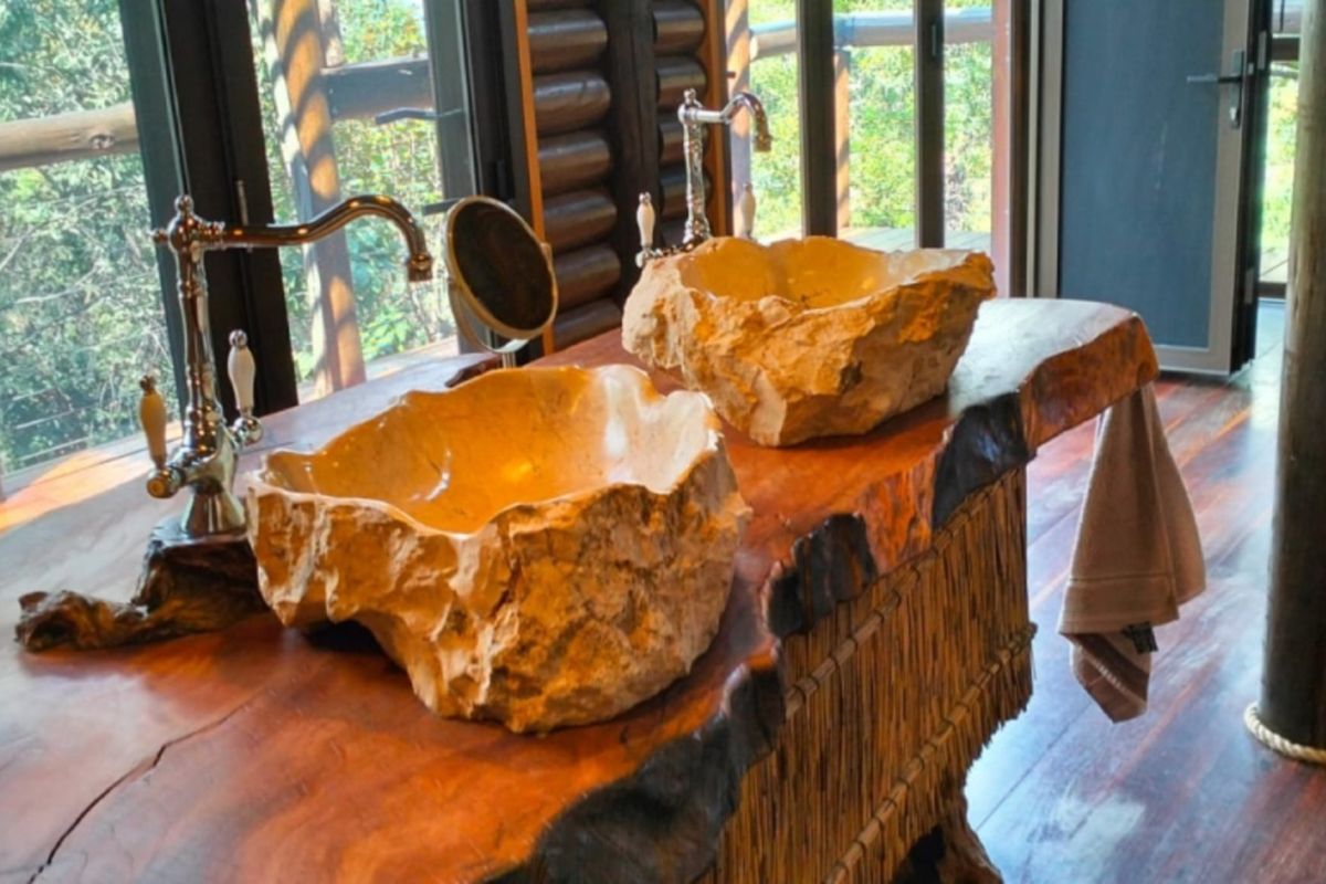 A luxury bathroom featuring a custom live-edge wooden vanity with dual natural stone vessel sinks and vintage-style chrome fixtures, overlooking a forest view.