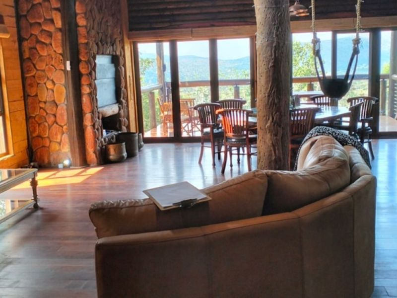 Spacious open-plan living area featuring rustic log walls, a stone fireplace, timber flooring, and large glass doors opening to a scenic outdoor deck.