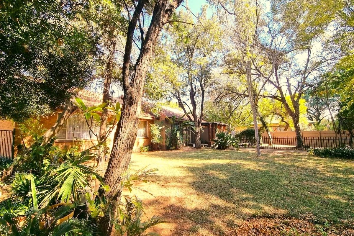 A spacious rear garden featuring a brick house facade, mature trees, and secure metal perimeter fencing.