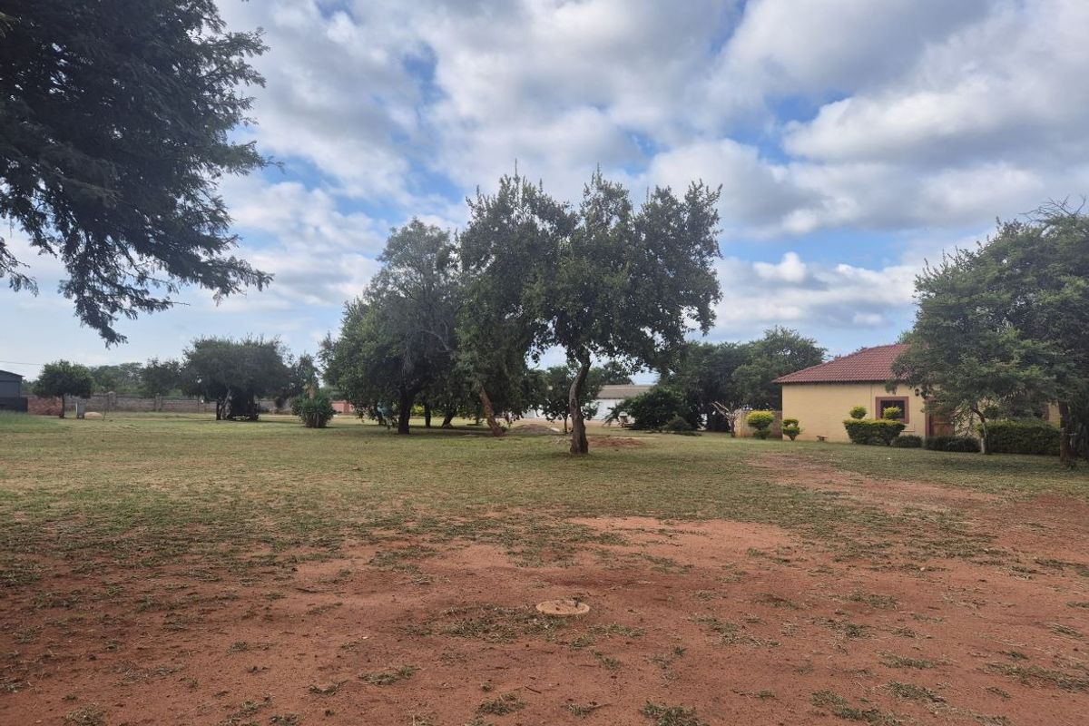 A spacious, open residential plot featuring established trees, a large grassed area, and a partial view of an existing structure.