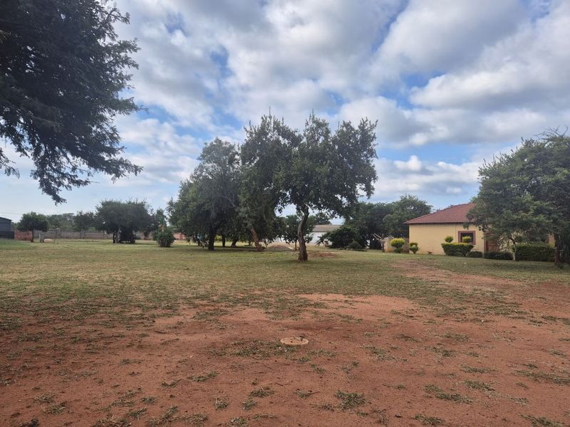 A spacious, open residential plot featuring established trees, a large grassed area, and a partial view of an existing structure.