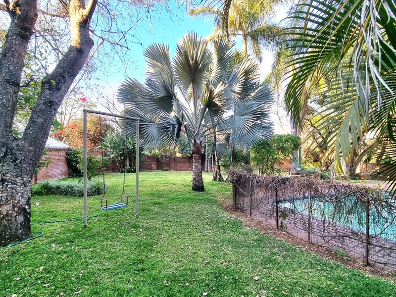 A spacious garden area featuring a swimming pool with a metal safety fence and established trees, enclosed by a brick boundary wall.