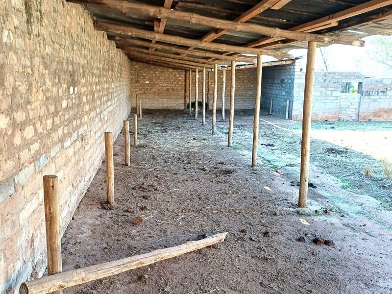 An open-sided farm utility structure featuring a corrugated metal roof supported by timber posts and an exposed brick wall with a dirt floor.