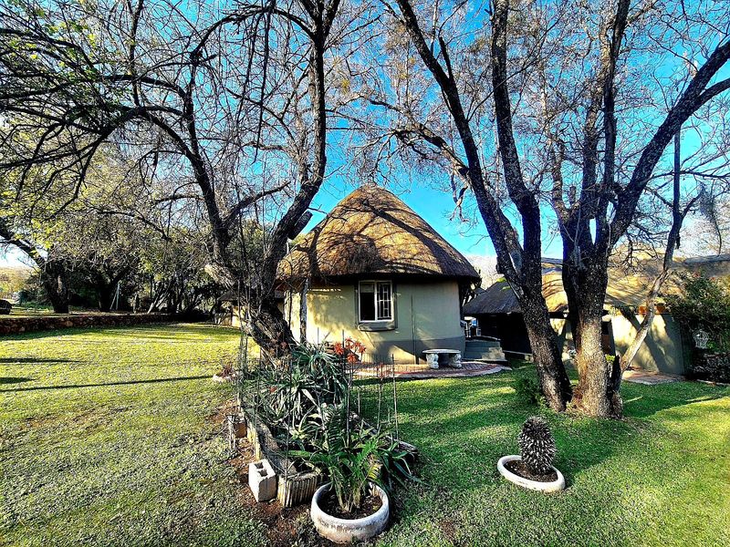 A charming thatched-roof cottage set within a spacious, well-maintained garden with mature trees and a paved patio area.