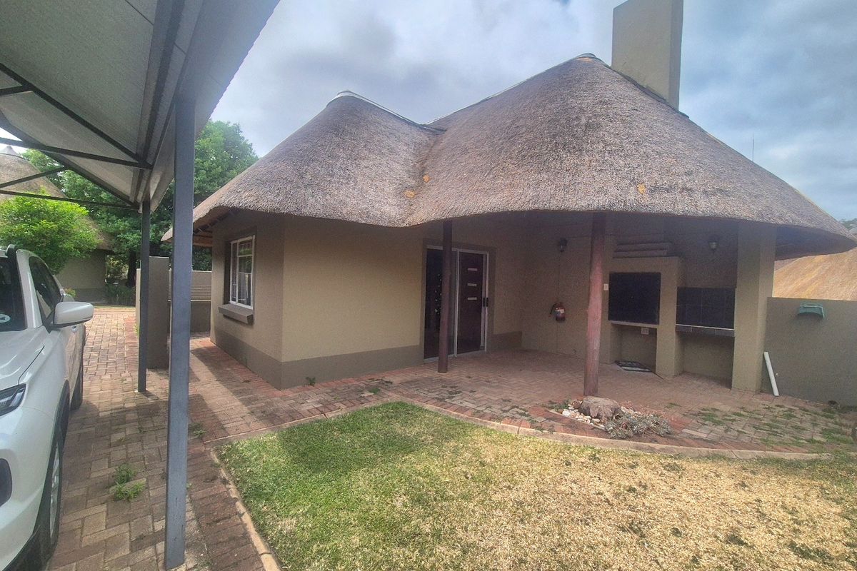 Rear view of a thatch-roof house featuring a paved patio area with a built-in braai and an adjacent carport.
