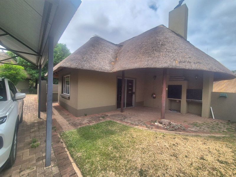 Rear view of a thatch-roof house featuring a paved patio area with a built-in braai and an adjacent carport.
